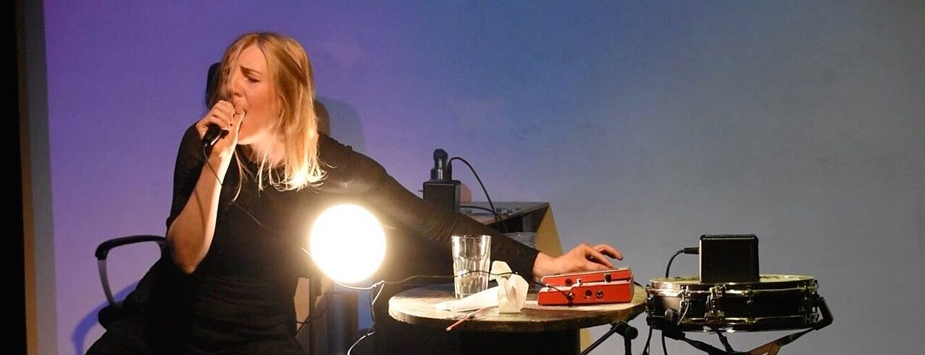 Person mit langen blonden Haaren sitzt an einem runden Tisch und singt in ein Mikrofon