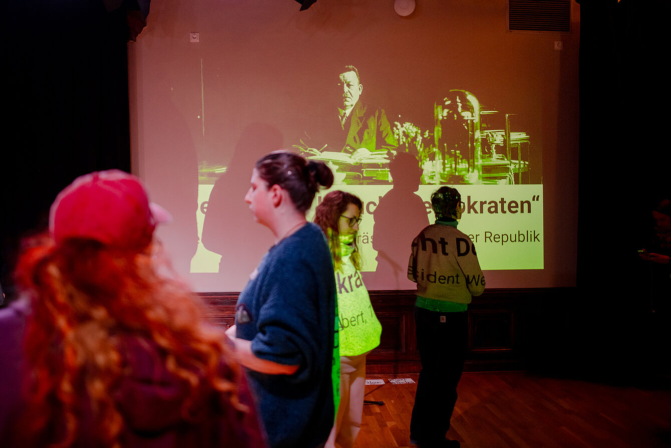 Mehrere Personen stehen vor einer großen Leinwand mit einem historischen Foto und Text, darunter eine Person am Tisch und der Schriftzug 'Demokraten'.