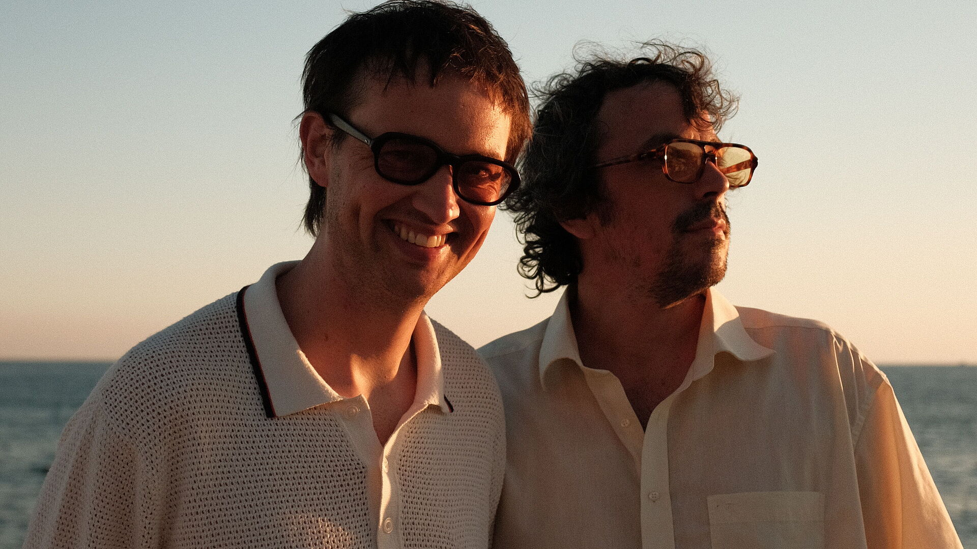 zwei männlich gelesene Personen stehen im Sonnenuntergang beim Meer mit Sonnenbrillen und grinsen in die Kamera