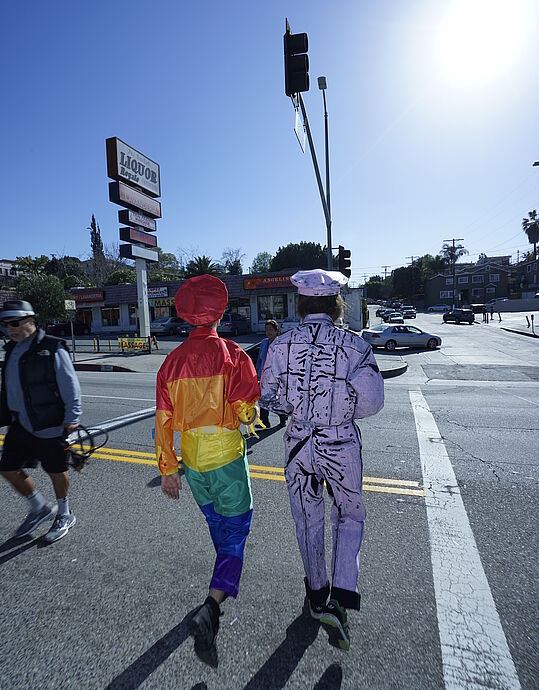 Zwei Personen in Uniformen bestehend aus Regenbogenstoff und Subwaymap-Stoff überqueren eine amerikanische Kreuzung.