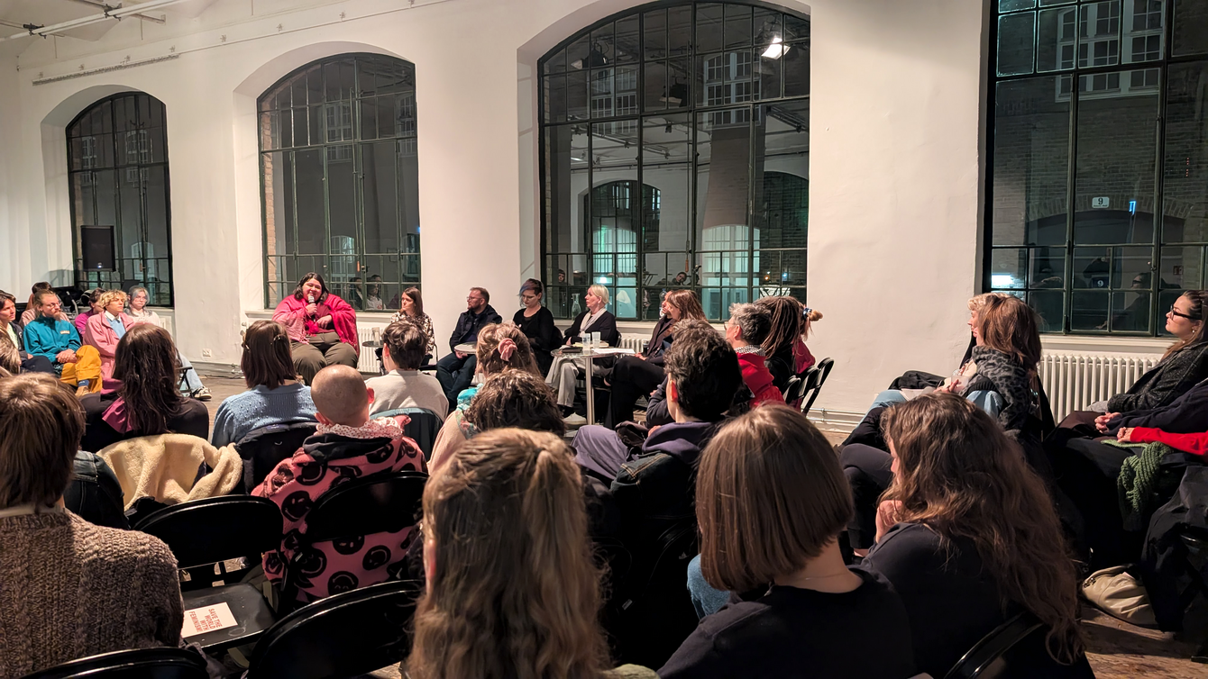 Gruppe von Menschen sitzt in einem Raum mit großen Fenstern und hört einer sprechenden Person zu