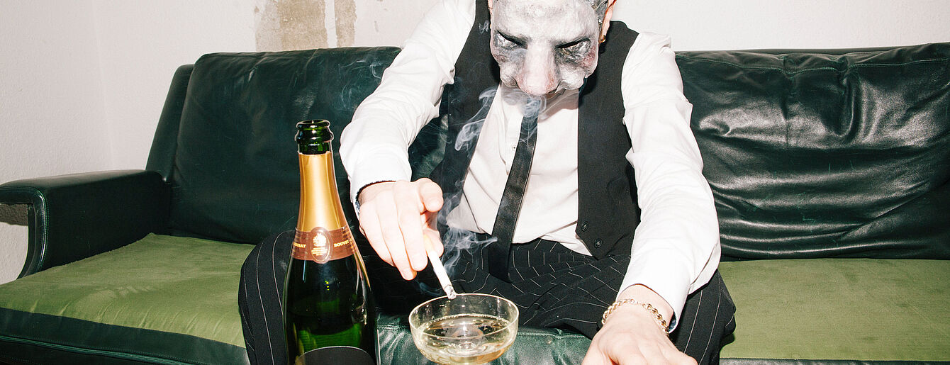 ein Mann mit Hasenmaske sitzt auf einem Sofa und ascht seine Zigarete in einen Champagnekelch