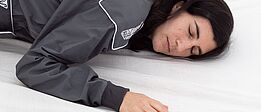 eine weiblich gelesene Person in einem dunkelgrauen Overall und mit dunklen Haaren, liegt auf einer Matratze mit weißem Leintuch. Die Augen sind geschlossen.