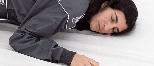 eine weiblich gelesene Person in einem dunkelgrauen Overall und mit dunklen Haaren, liegt auf einer Matratze mit weißem Leintuch. Die Augen sind geschlossen.