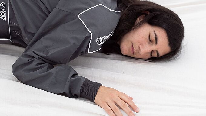 eine weiblich gelesene Person in einem dunkelgrauen Overall und mit dunklen Haaren, liegt auf einer Matratze mit weißem Leintuch. Die Augen sind geschlossen.