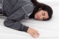 eine weiblich gelesene Person in einem dunkelgrauen Overall und mit dunklen Haaren, liegt auf einer Matratze mit weißem Leintuch. Die Augen sind geschlossen.
