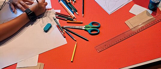 Kinder zeichnen an einem Tisch mit verstreuten Buntstiften, Schere, Linealen und Radiergummi