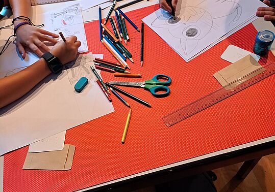 Kinder zeichnen an einem Tisch mit verstreuten Buntstiften, Schere, Linealen und Radiergummi