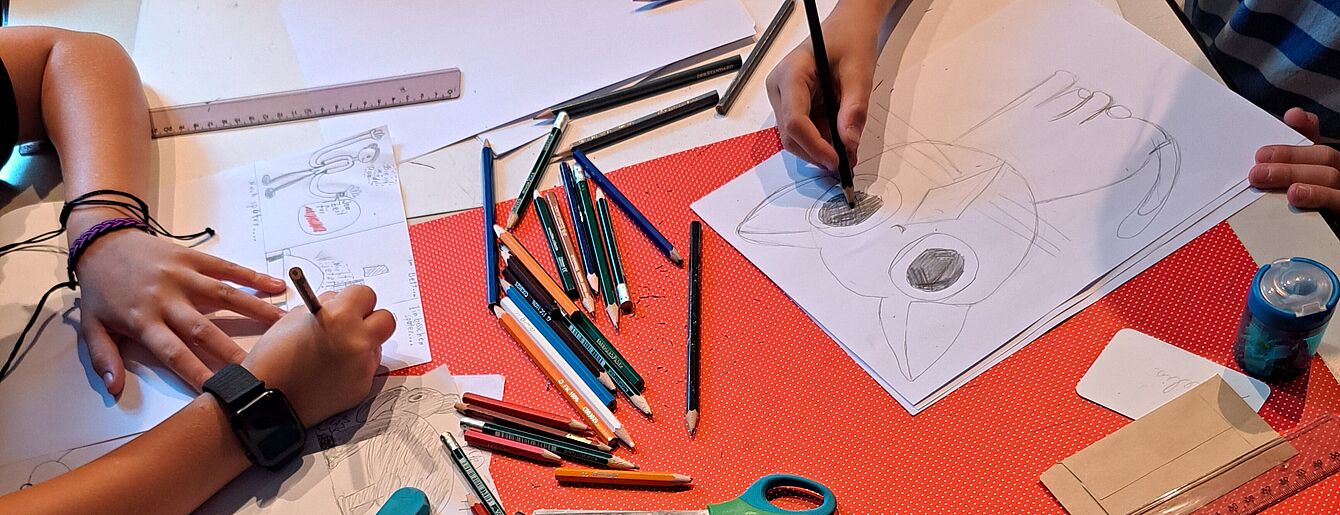 Kinder zeichnen an einem Tisch mit verstreuten Buntstiften, Schere, Linealen und Radiergummi