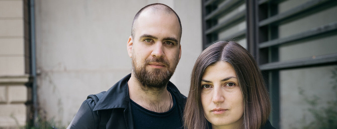 Porträt von Natalia und Yuri vor einem Gebäude mit Fenster und grauer Fassade
