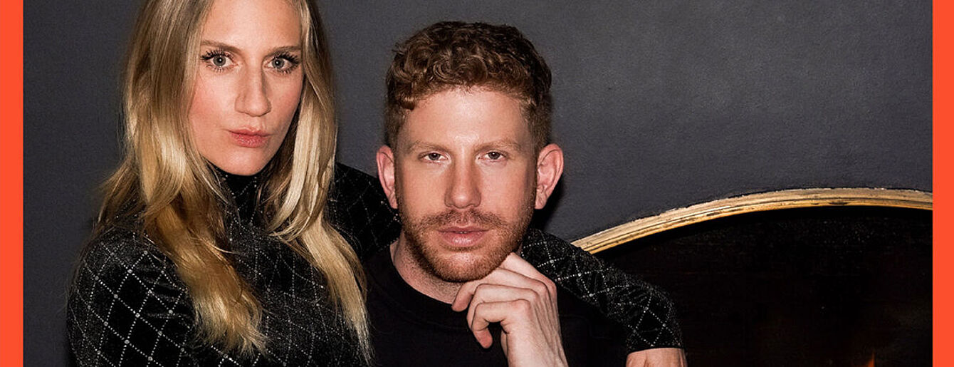 Zwei Personen sitzen vor einer dunklen Wand, die Person links hat lange blonde Haare und trägt ein kariertes Oberteil, der Mann hat kurze lockige Haare und stützt das Kinn auf die Hand