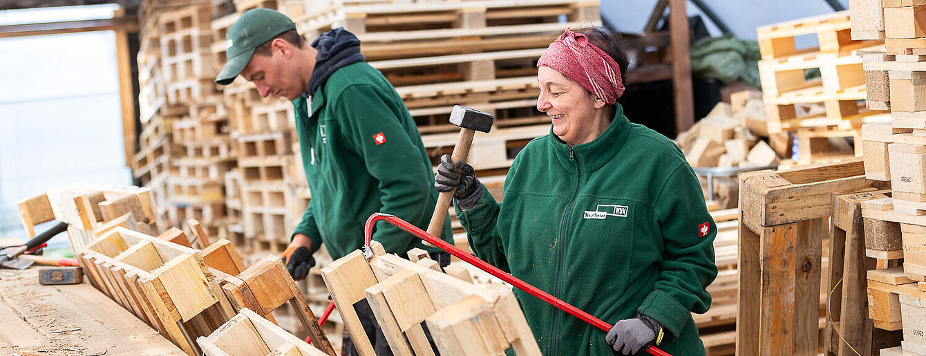 Zwei Personen in grünen Arbeitsjacken bearbeiten Holzpaletten in einer Werkstatt von WUK re.cycle