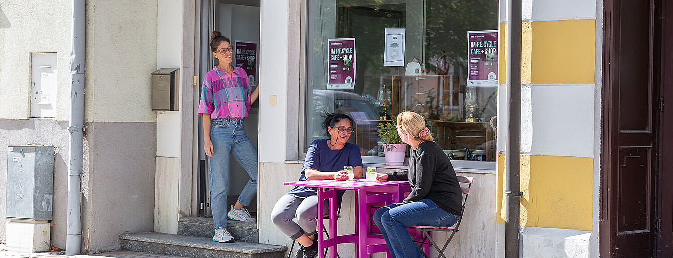 zwei Personen sitzen an einem kleinen lila Tisch vor einem Café, eine weitere Person steht in der Tür. Bunte Girlande hängt im Fenster