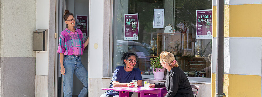 Foto: Susanne Senekowitsch zwei Personen sitzen an einem kleinen lila Tisch vor einem Café, eine weitere Person steht in der Tür. Bunte Girlande hängt im Fenster