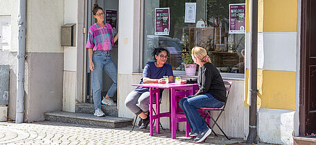 Foto: Susanne Senekowitsch wei Personen sitzen an einem kleinen lila Tisch vor einem Café, eine weitere Person steht in der Tür. Bunte Girlande hängt im Fenster