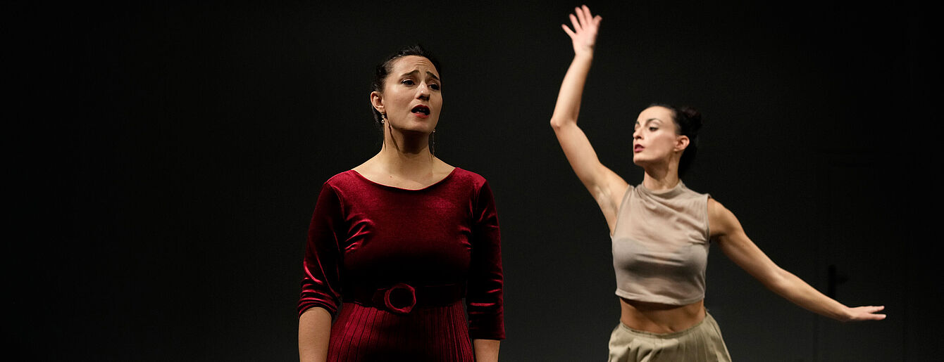 Zwei Personen bei einer Tanzperformance auf dunkler Bühne, eine im Vordergrund mit rotem Samtkleid