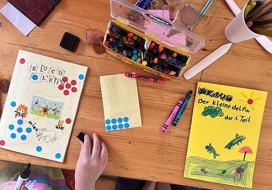 Kinderhand malt auf einem Tisch mit Buntstiften und Wasserfarben an selbstgebundenen Büchern mit bunten Punkten und Zeichnungen