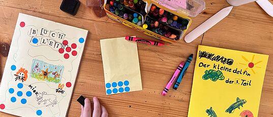 Kinderhand malt auf einem Tisch mit Buntstiften und Wasserfarben an selbstgebundenen Büchern mit bunten Punkten und Zeichnungen