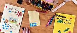 Kinderhand malt auf einem Tisch mit Buntstiften und Wasserfarben an selbstgebundenen Büchern mit bunten Punkten und Zeichnungen