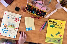 Kinderhand malt auf einem Tisch mit Buntstiften und Wasserfarben an selbstgebundenen Büchern mit bunten Punkten und Zeichnungen