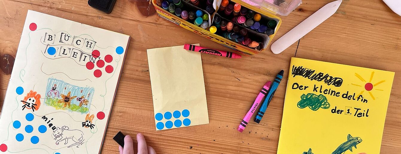Kinderhand malt auf einem Tisch mit Buntstiften und Wasserfarben an selbstgebundenen Büchern mit bunten Punkten und Zeichnungen