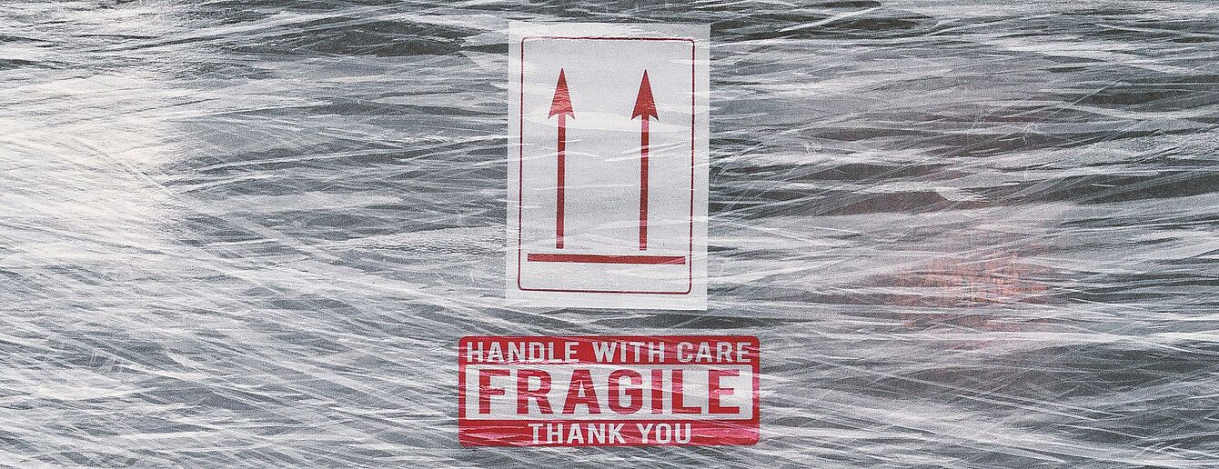Verpackung mit Aufklebern 'Handle with care Fragile Thank you' und zwei nach oben zeigenden Pfeilen auf transparentem Stretchfolie