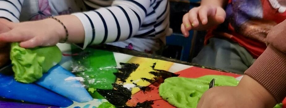 Kinderhände formen grüne Knetmasse auf einem bunt bemalten Tisch mit Regenbogenfarben