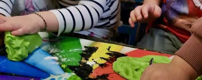Kinderhände formen grüne Knetmasse auf einem bunt bemalten Tisch mit Regenbogenfarben