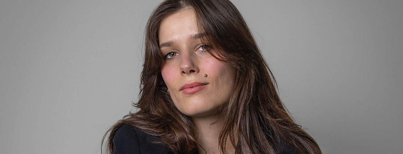 Tara-Louise_Wittwer_WUK_2025_(c)_LewisJones Porträt von Tara Louise Wittwer mit langen braunen Haaren und schwarzem Oberteil vor grauem Hintergrund