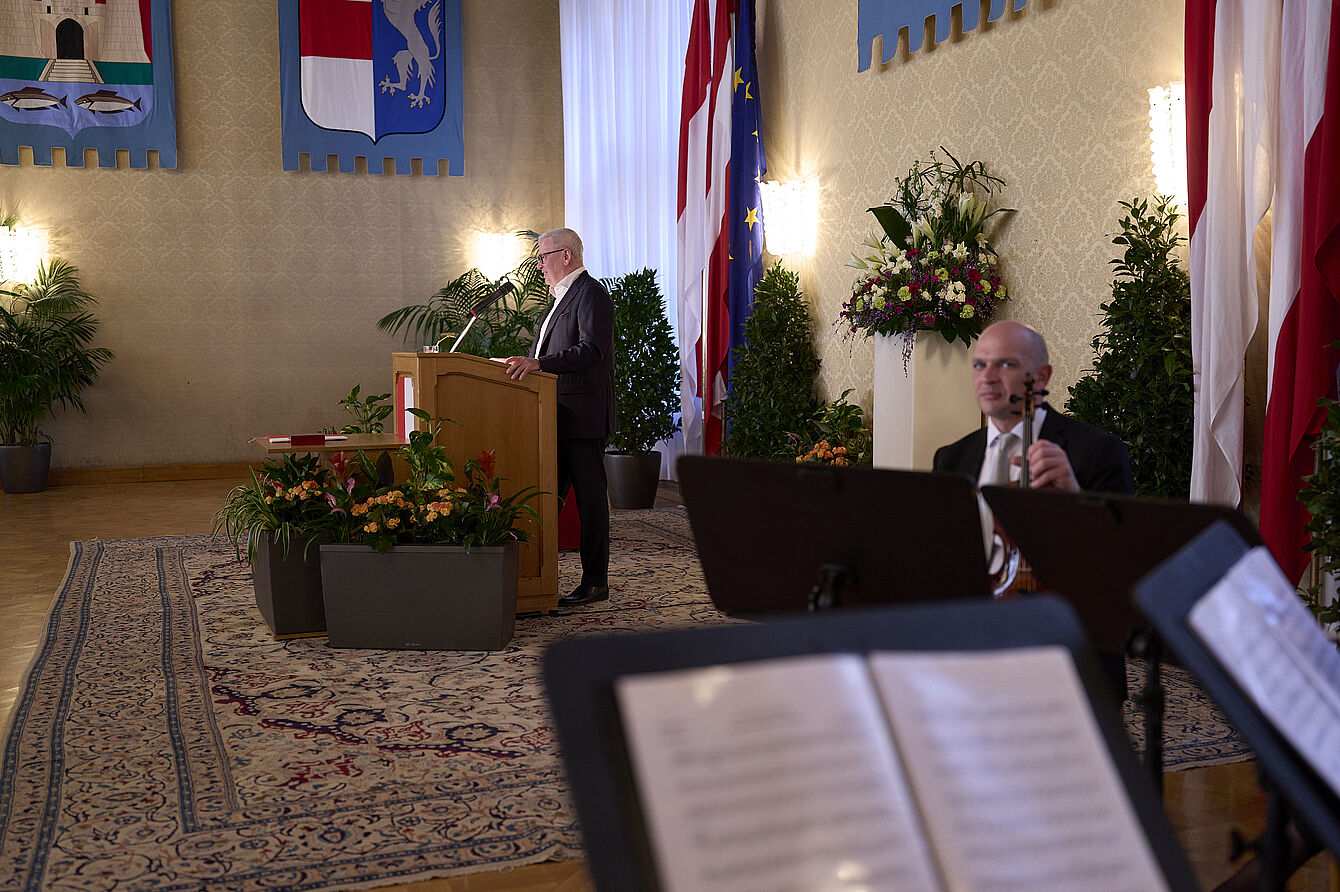 Mann steht an einem Rednerpult in einem festlich dekorierten Raum mit Fahnen, Pflanzen und Teppich, im Vordergrund unscharfe Notenblätter und ein Musiker mit Klarinette