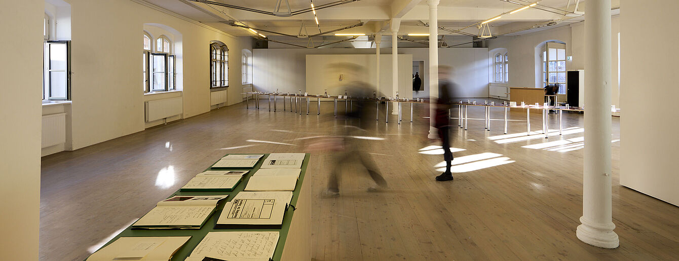 Large exhibition room of kex with wooden floor and tall windows. A long table displays several guest books. Two blurred figures walk through the space.