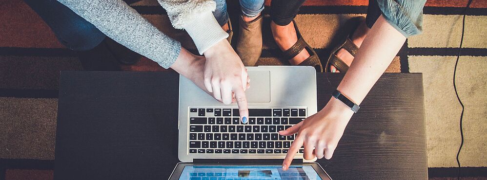 Zwei Personen zeigen mit Fingern auf den Bildschirm eines Laptops auf einem Tisch