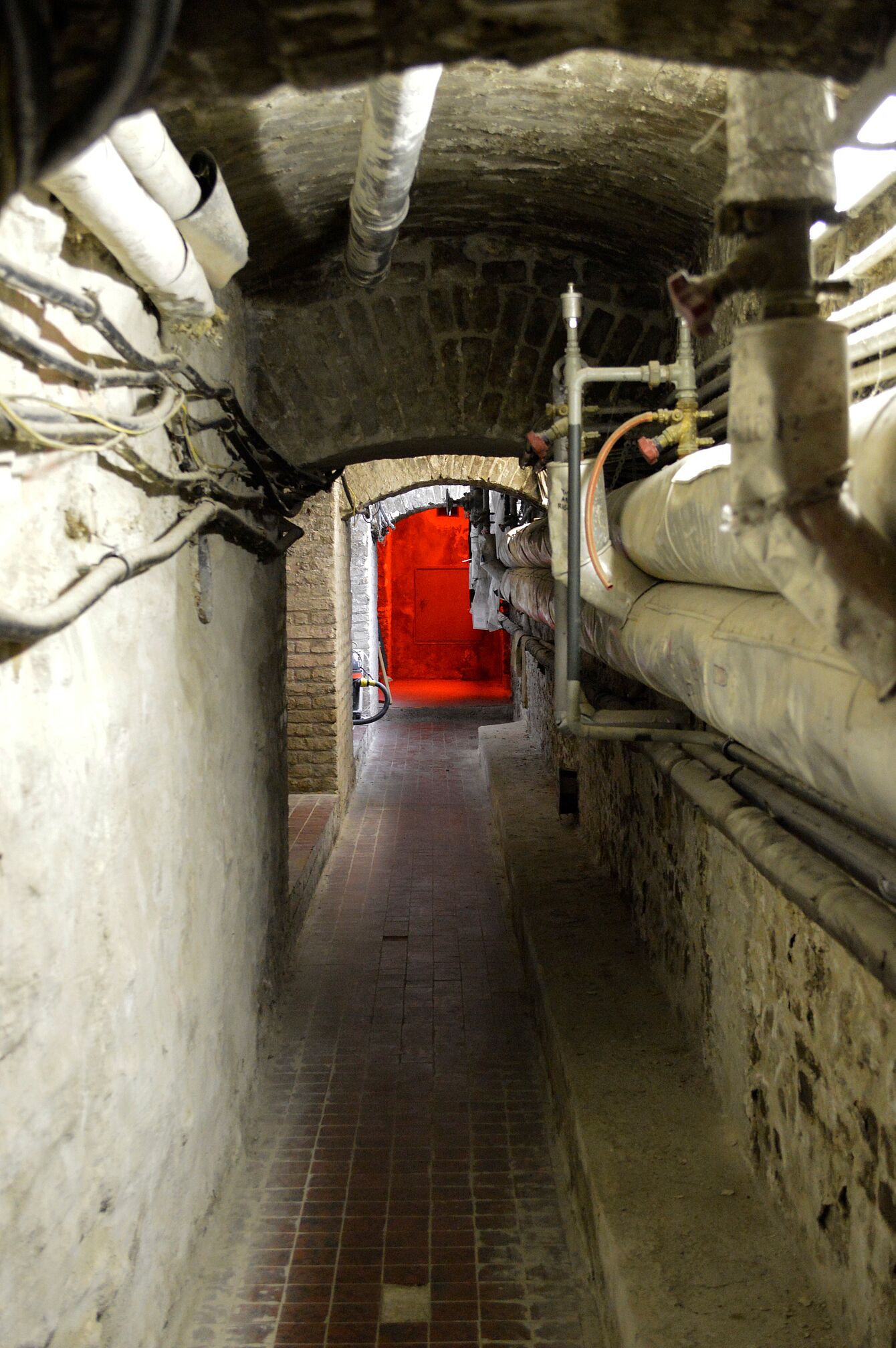 Schmaler Gang mit Backsteinboden und sichtbaren Rohren an den Wänden führt zu einem Durchgang mit rotem Licht im Hintergrund