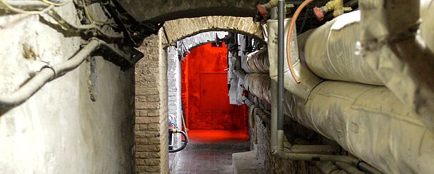 Schmaler Gang mit Backsteinboden und sichtbaren Rohren an den Wänden führt zu einem Durchgang mit rotem Licht im Hintergrund