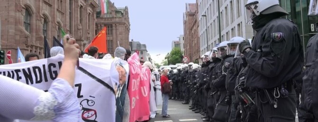 Das Bild eine Straße, rechts eine Reihe von Polizist*innen und links eine Reihe Demonstrant*innen mit Transparenten