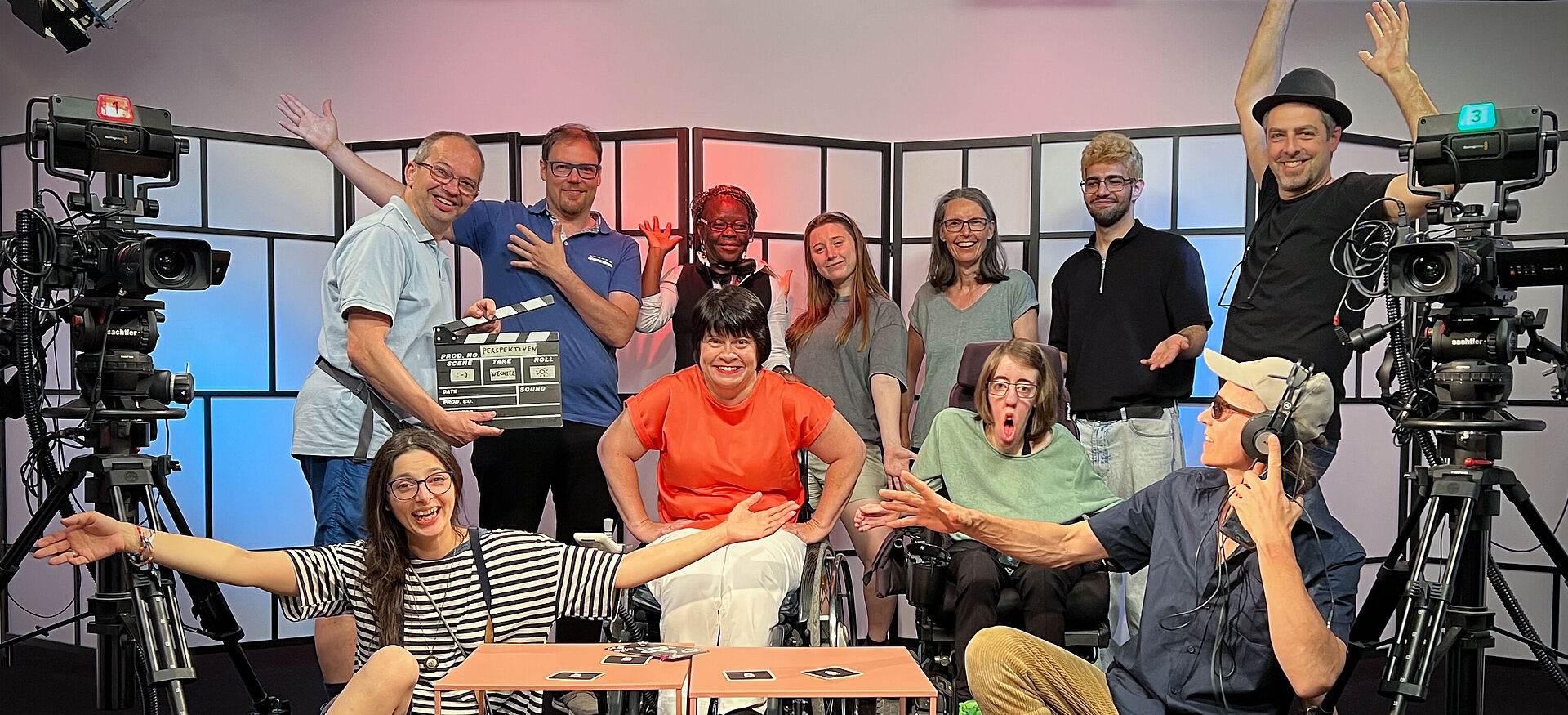 11 Personen sitzen, zwei davon im Rollstuhl, und stehen in einem TV-Studio und blicken in die Kamera