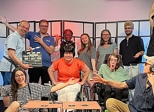 11 Personen sitzen, zwei davon im Rollstuhl, und stehen in einem TV-Studio und blicken in die Kamera