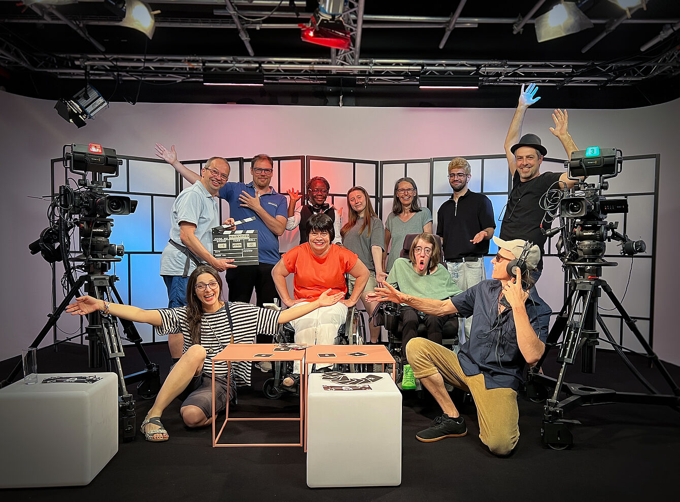11 Personen sitzen, zwei davon im Rollstuhl, und stehen in einem TV-Studio und blicken in die Kamera