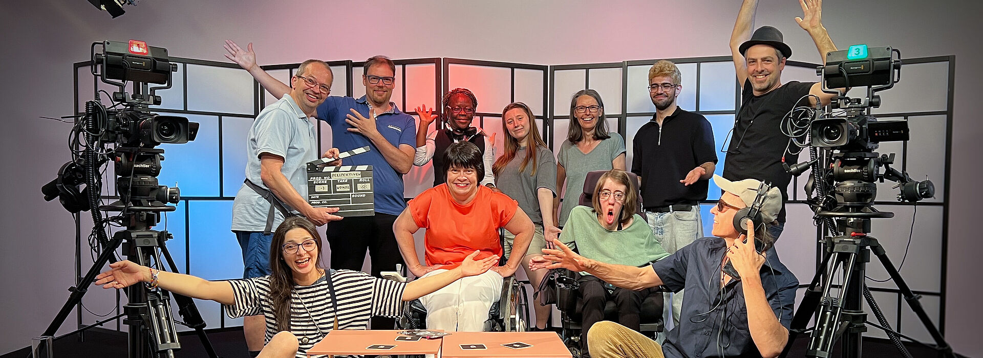 11 Personen sitzen, zwei davon im Rollstuhl, und stehen in einem TV-Studio und blicken in die Kamera