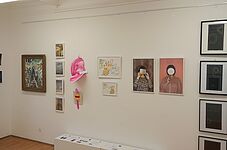 Galeriewand mit verschiedenen gerahmten Bildern, darunter zwei Fotografien von Personen mit verdecktem Gesicht und eine pinkfarbene Skulptur an der Wand
