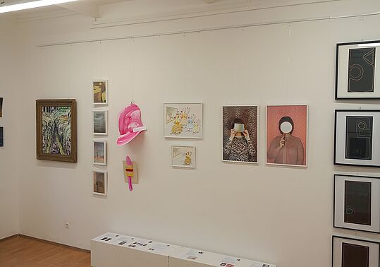 Galeriewand mit verschiedenen gerahmten Bildern, darunter zwei Fotografien von Personen mit verdecktem Gesicht und eine pinkfarbene Skulptur an der Wand