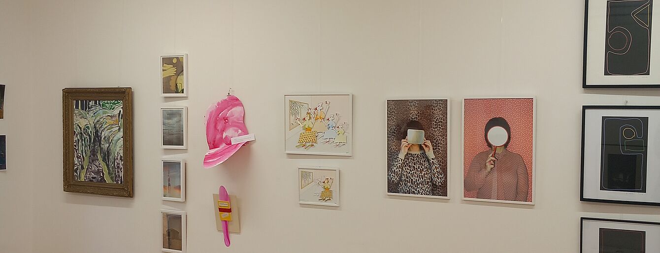 Galeriewand mit verschiedenen gerahmten Bildern, darunter zwei Fotografien von Personen mit verdecktem Gesicht und eine pinkfarbene Skulptur an der Wand