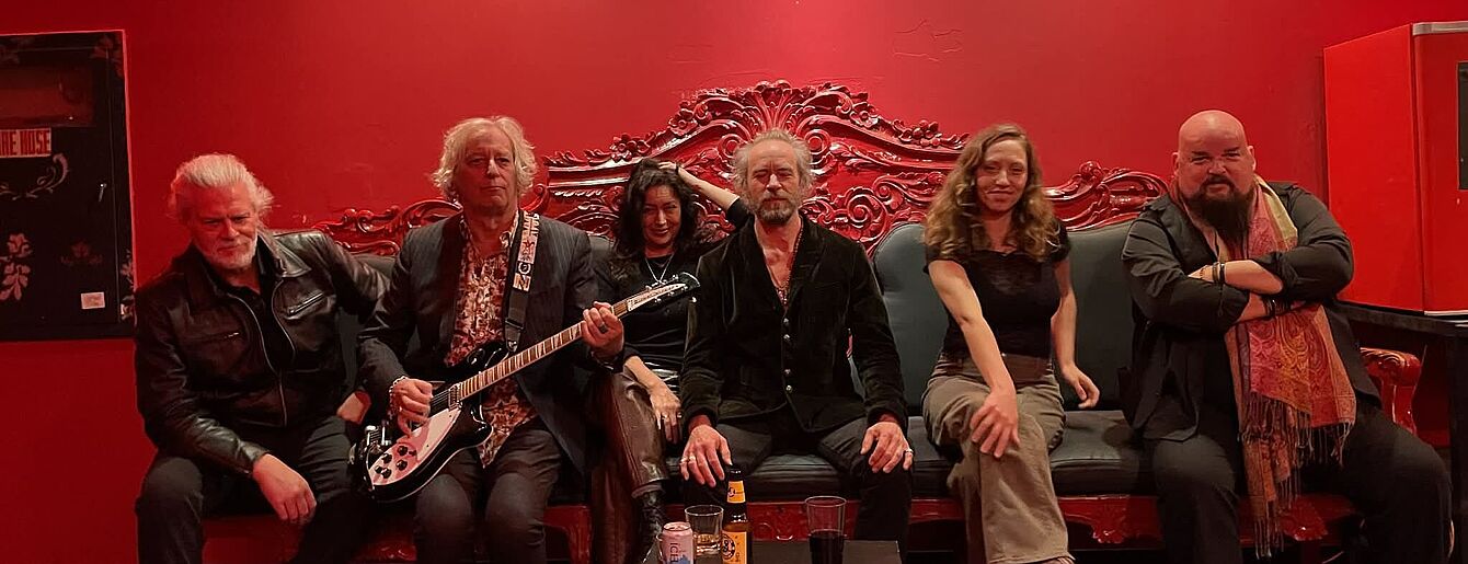 Sechs Personen sitzen nebeneinander auf einer Couch vor einer rot beleuchteten Wand, zwei von ihnen halten Gitarren, alle blicken frontal zur Kamera