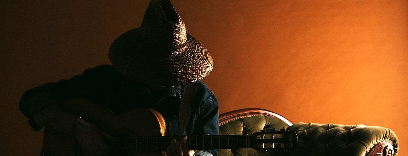 Person mit Strohhut spielt Gitarre auf grünem Sofa vor orangefarbener Wand 