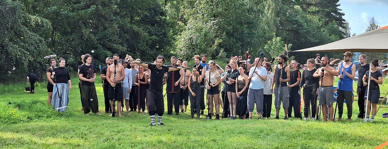 Eine Gruppe von Mernschen auf einer grünen Wiese, alle mit langen Stöcken, notwendig für das Training von Fow Arts.