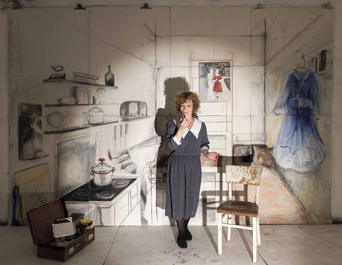 Frau in gepunktetem Kleid steht in einem Raum mit skizzenhafter Kücheneinrichtung an der Wand und einem blauen Kleid, das an der Wand hängt