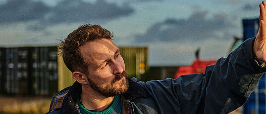 Florian Künstler steht zwischen Containern und blickt gegen die Sonne, mit seiner Hand versucht er das Sonnenlicht aus seinem Gesicht zu halten.