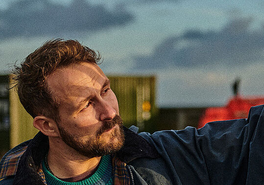 Florian Künstler steht zwischen Containern und blickt gegen die Sonne, mit seiner Hand versucht er das Sonnenlicht aus seinem Gesicht zu halten.