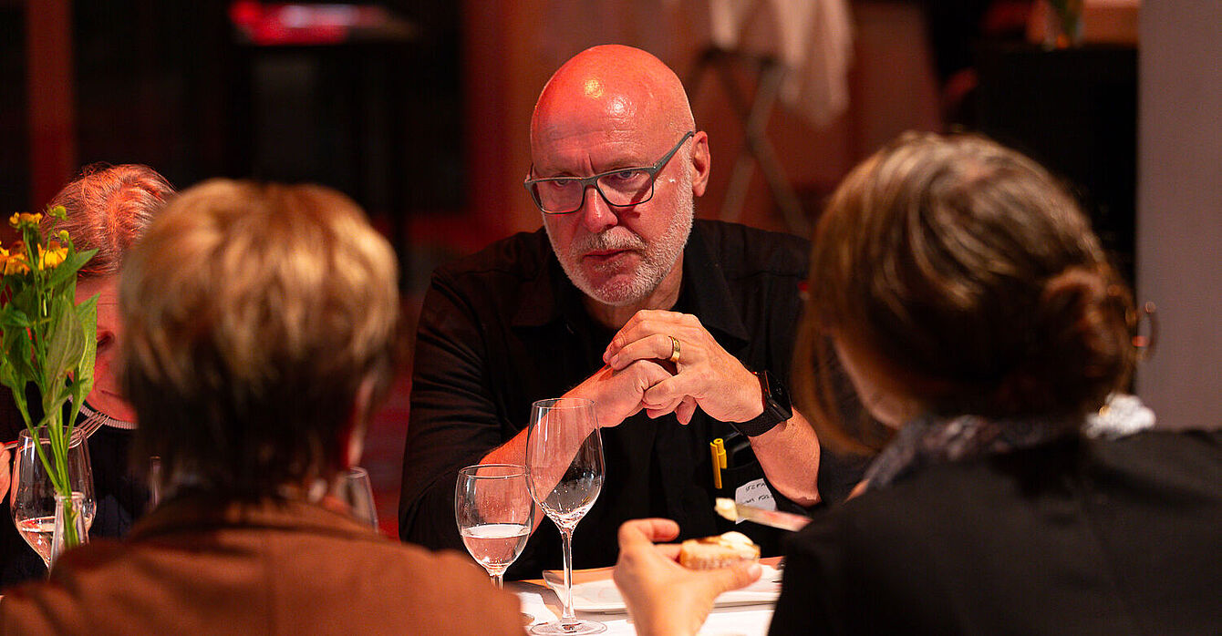 Foto: Susanne Senekowitsch Drei Personen sitzen an einem Tisch mit weißen Tischdecke, zwei Frauen von hinten, ein Mann mit schwarzem Hemd von vorne, auf dem Tisch Weingläser und eine Blume in Vase.