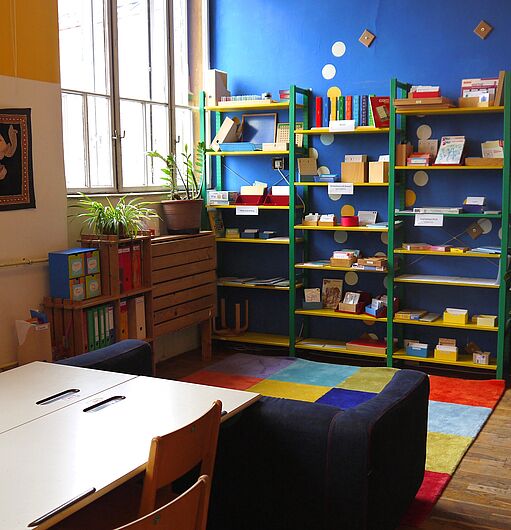 Bunt eingerichteter Raum mit blauer Wand, Regalen voller Bücher und Gegenstände, buntem Teppich, mehreren Tischen und Stühlen sowie Pflanzen am Fenster und an der Wand